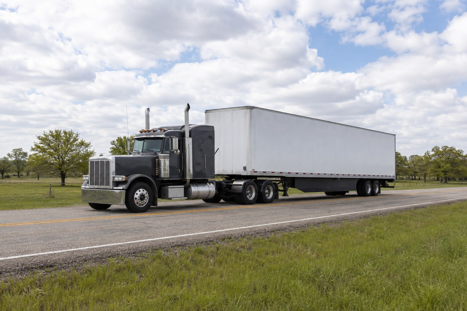 A charcoal semi-truck with a white trailer on a green Texas-style rural road